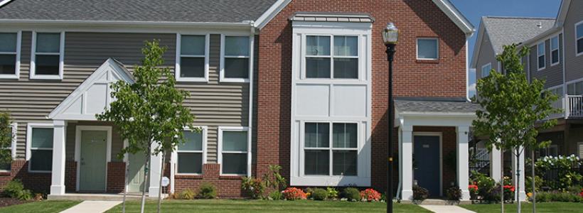 A row of two story town homes, the exterior is red, gray, and white.  Each has a covered patio and small lawn.